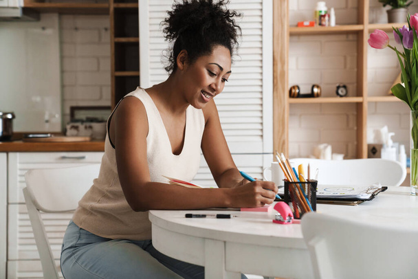 Black smiling woman working with papers while sitting at table in home kitchen - Foto, imagen