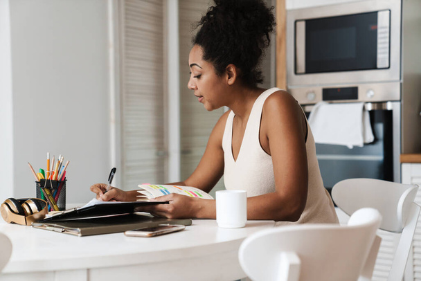 Чорна жінка працює з паперами, сидячи за столом на домашній кухні
 - Фото, зображення