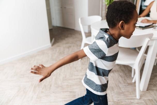Black boy running and making fun while her mother working at table in home - Photo, Image