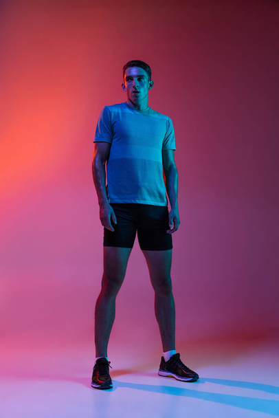 Portrat of Caucasian young sportive man in sportswear standing isolated on pink studio background with blue neon filter, light. Muscular male model. - Foto, Bild