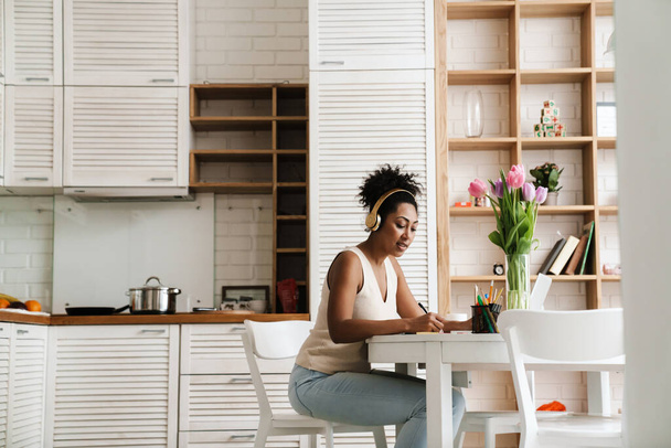 Black woman in headphones working with laptop while sitting at table in home kitchen - Foto, immagini