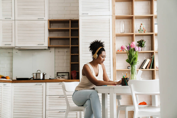 Black woman in headphones working with laptop while sitting at table in home kitchen - Fotoğraf, Görsel
