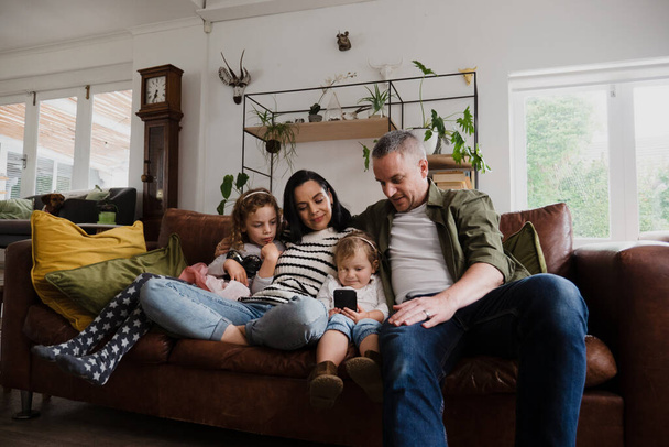 Szczęśliwa rodzina siedzi razem, podczas gdy dziecko trzyma smartfona, siedzi na kanapie w salonie. - Zdjęcie, obraz