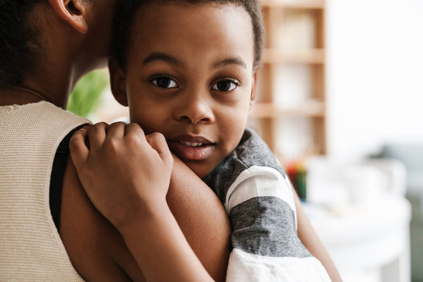 zwart elementaire jongen kijken naar camera terwijl knuffelen haar moeder thuis - Foto, afbeelding