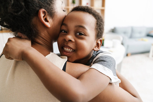 Black elementary boy smiling while hugging her mother at home - Foto, Imagen
