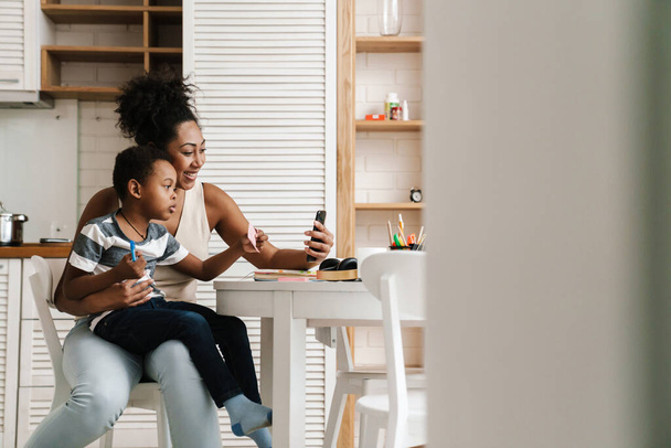 Černá usměvavá matka a syn pomocí mobilního telefonu, zatímco sedí spolu doma - Fotografie, Obrázek