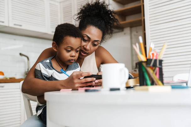 Black mother and son using mobile phone while sitting together at home - Fotoğraf, Görsel