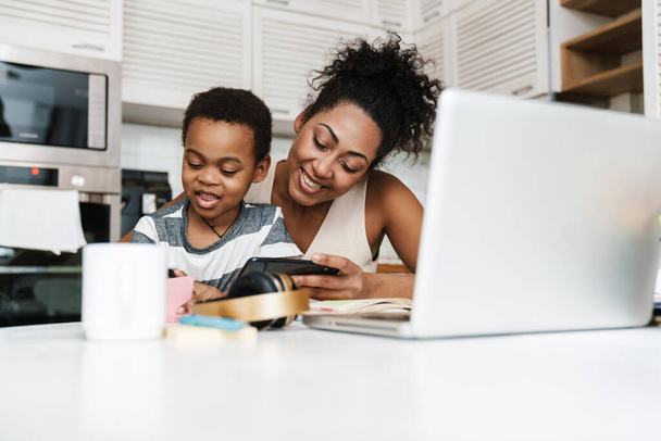 Black smiling mother and son using mobile phone and laptop while sitting together at home - Fotoğraf, Görsel