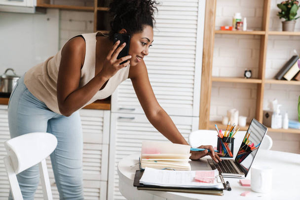 Black smiling woman talking on cellphone while working with laptop at home - Foto, Bild
