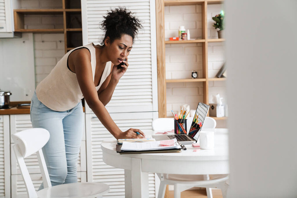 Black puzzled woman talking on cellphone while working with laptop at home - Photo, Image