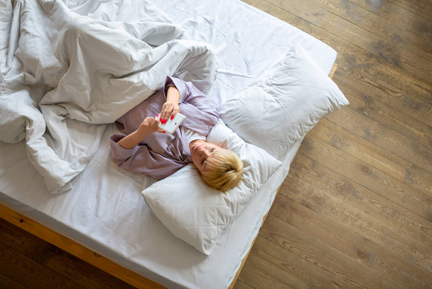 Κάτοψη της χαμογελαστή ξανθιά γυναίκα ξαπλωμένη στο κρεβάτι με smartphone. - Φωτογραφία, εικόνα