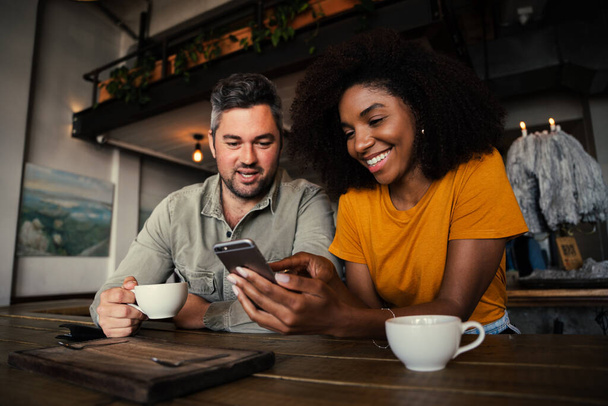 Happy couple smiling looking at cute pictures on smartphone while enjoying cup of coffee in trendy cafe  - Foto, Imagen