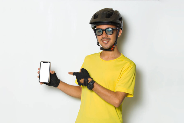 young handsome man wearing cyclist helmet hold mobile phone with blank empty screen isolated on white background - Photo, Image