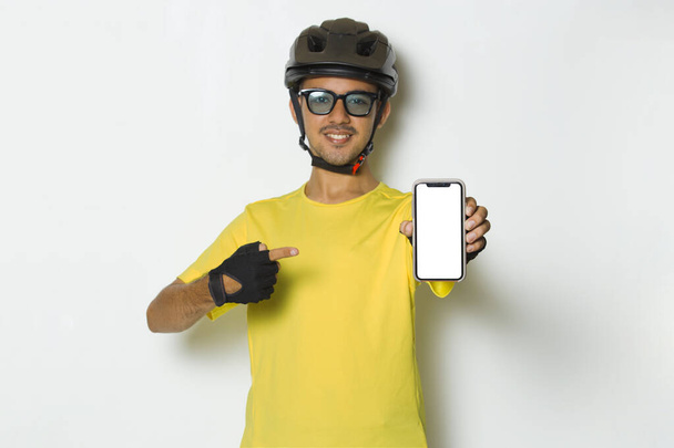 fiatal jóképű férfi visel kerékpáros sisak tartsa mobiltelefon üres képernyő elszigetelt fehér háttér - Fotó, kép
