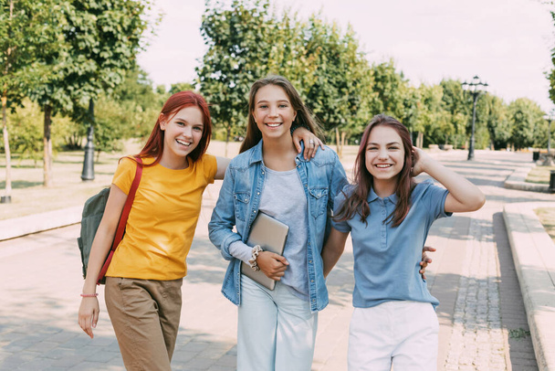 Троє дівчат-підлітків ходять з планшетом і рюкзаком вздовж алеї парку до школи. Концепція підготовки та виховання. Назад до школи
 - Фото, зображення