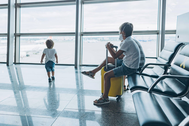Tyhjän lentokentän terminaalin lapset odottavat lähtöä ikkunasta. Pikkupojat, joilla on suojaava kasvonaamio, istuvat aulassa odottamassa lentoa. Perhematkan käsite - Valokuva, kuva