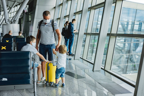 空港で黄色のスーツケースと保護医療面マスクの父と息子。飛行機の窓から外を見る子供たち。家族の出発ゲートを待っている。家族旅行と休暇のコンセプト. - 写真・画像
