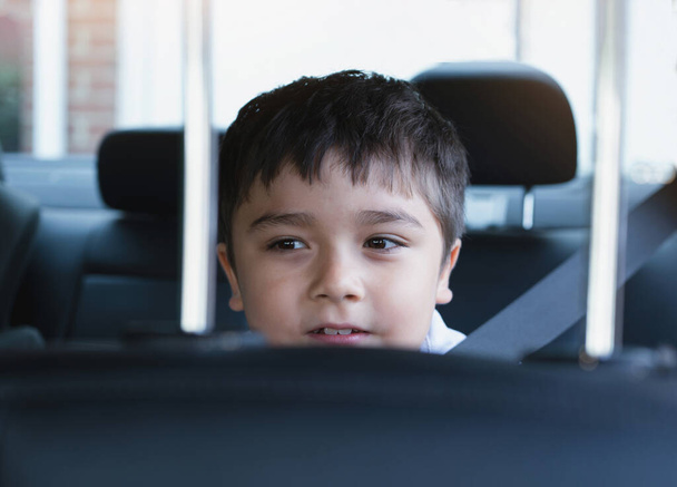 Primer plano de la cara de niño lindo sentado en el asiento de seguridad del coche mirando hacia fuera con la cara sonriente, Niño sentado en el asiento trasero del pasajero con un cinturón de seguridad, Niño de la escuela viajando a la escuela en coche.Volver a la escuela - Foto, Imagen
