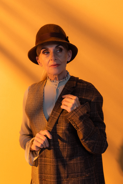 Senior woman in vintage clothes and hat holding jacket on orange background  - Фото, зображення