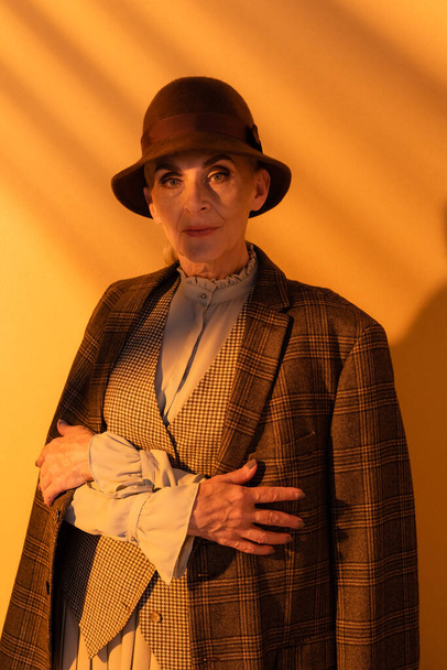 Senior woman in hat and plaid jacket looking at camera on orange background  - Fotoğraf, Görsel