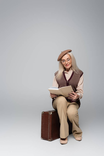Elderly woman in beret reading book on retro suitcase on grey background  - Photo, image