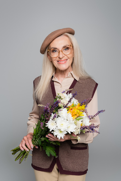 Stylish senior woman smiling while holding flowers isolated on grey  - Foto, immagini