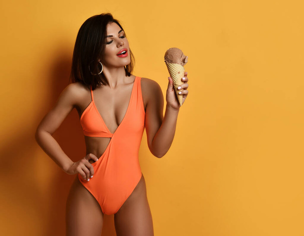Young smiling sexy pretty lady in orange swimwear bikini standing and holding chocolate ice cream in hand - Photo, Image