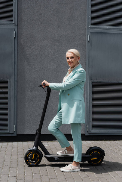 Glimlachende oudere zakenvrouw in de buurt van elektrische kick scooter  - Foto, afbeelding