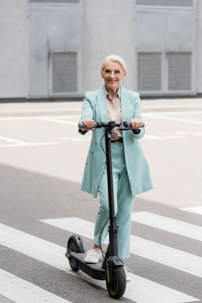 Mulher de negócios sênior alegre que está perto de scooter de chute elétrico  - Foto, Imagem