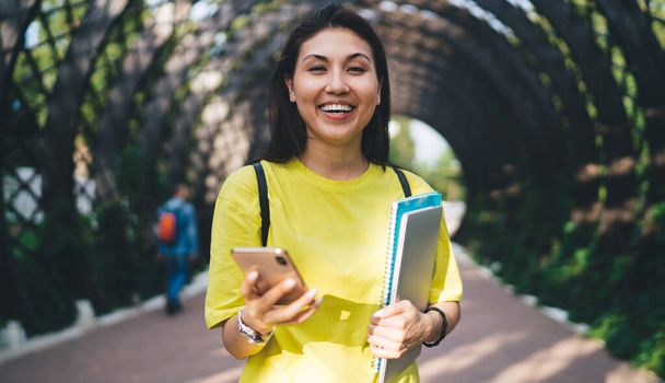 Half length portrait of cheerful asian woman in casual wear laughing at funny messages standing on street with cellular, happy young 20s female looking at camera using smartphone sending emails - Zdjęcie, obraz