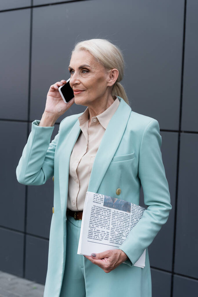 Senior businesswoman holding newspaper and talking on smartphone near building outdoors - Zdjęcie, obraz