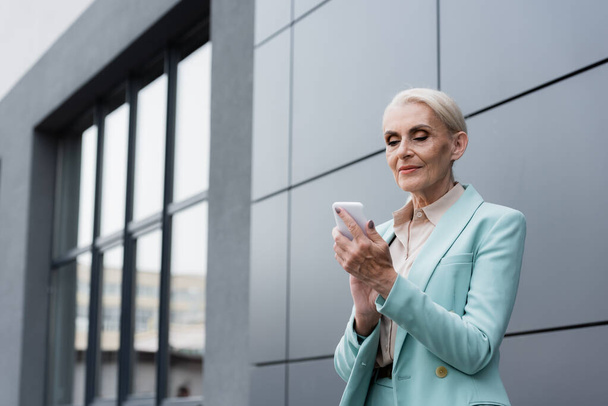 Elderly businesswoman using cellphone while standing near building outdoors - Photo, Image