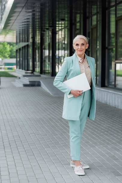 Senior businesswoman holding laptop and looking at camera near building outdoors - Foto, afbeelding
