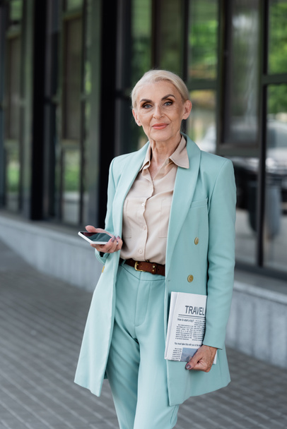 Senior businesswoman holding smartphone and newspaper outdoors  - Fotoğraf, Görsel