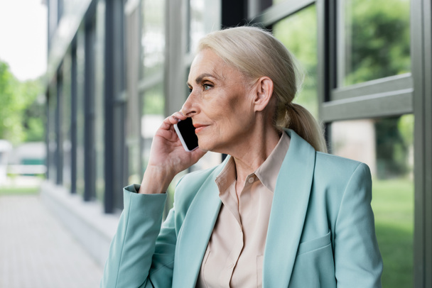 Side view of senior businesswoman talking on mobile phone near blurred building  - Fotoğraf, Görsel