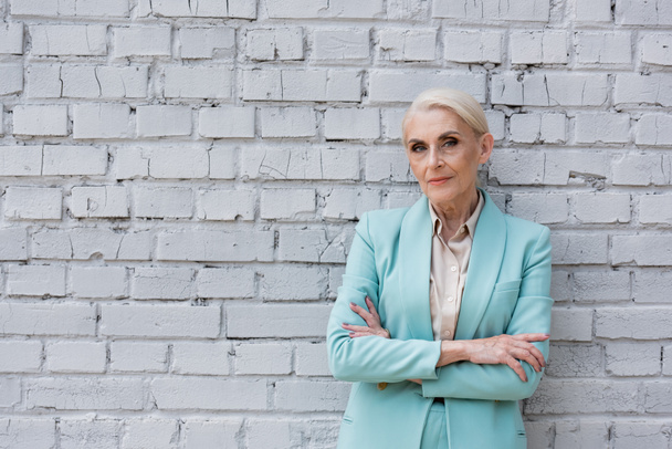 senior confident businesswoman standing with crossed arms near white brick wall - 写真・画像