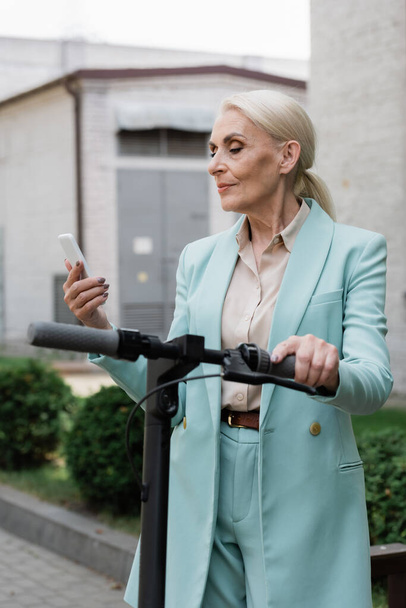 idosa empresária em blazer azul olhando para o telefone móvel perto de scooter chute elétrico - Foto, Imagem