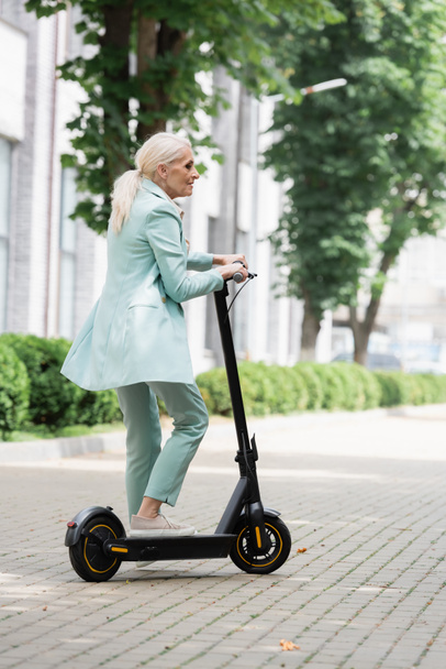 senior businesswoman in blue suit standing with electric kick scooter on city street - Fotoğraf, Görsel