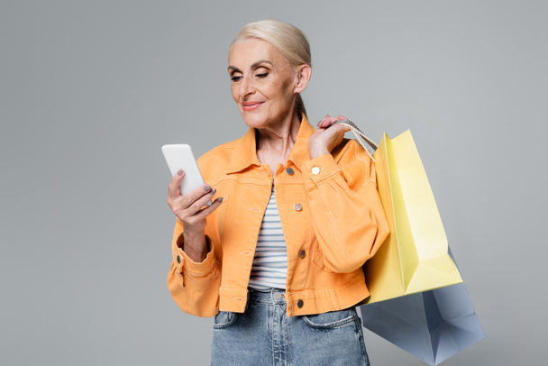 mujer mayor en chaqueta naranja con smartphone y bolsas de compras aisladas en gris - Foto, imagen