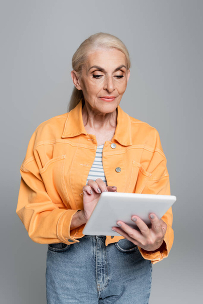 senior woman in orange jacket using digital tablet isolated on grey - Fotó, kép