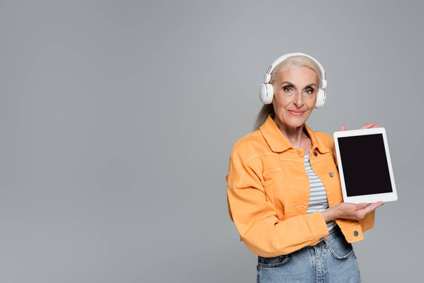 trendy senior woman in headphones showing digital tablet with blank screen isolated on grey - Fotó, kép