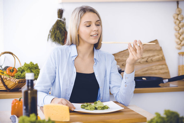 An attractive young blond woman tastes a new recipe for a delicious salad mix while sitting at the table in the kitchen. - Foto, afbeelding
