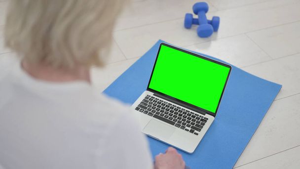 Mujer vieja usando el ordenador portátil con la pantalla verde en la estera del ejercicio - Foto, Imagen