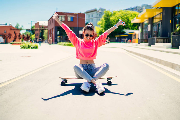 Carefree female skater 20s enjoying freedom youth lifestyle sitting at girlish skateboard and raising hands in air, happy woman dressed in trendy streetwear spending day for active board hobby - Фото, изображение