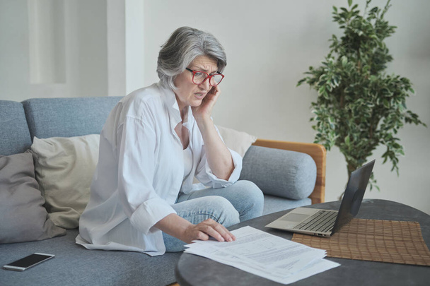 Пожилая женщина использует ноутбук, рассчитывая свои сбережения и разочаровываясь в долгах и огромных счетах. - Фото, изображение