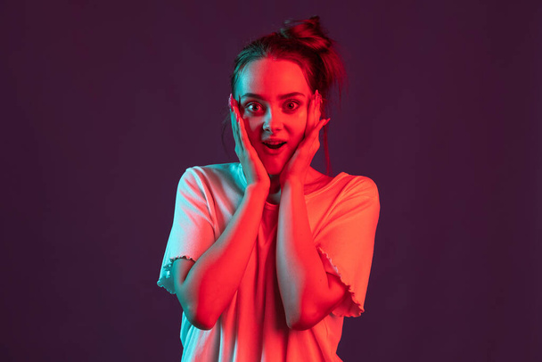 Portrait of beautiful girl in casual clothes isolated on dark purple studio background in red neon light, filter. Concept of human emotions, facial expression. - Foto, imagen