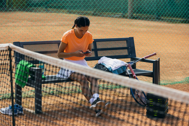 Μία νεαρή αθλήτρια, επαγγελματίας τενίστρια με τηλέφωνο στο γήπεδο τένις σε φωτεινή καλοκαιρινή μέρα. - Φωτογραφία, εικόνα