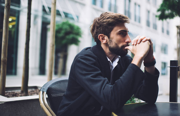 Thoughtful bearded male in dark jacket and light shirt thinking while sitting at empty table in outdoor cafe in summer day - 写真・画像