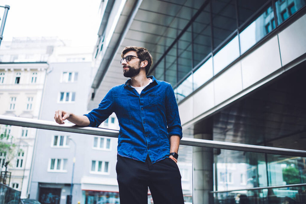 Low angle of peaceful young bearded male in eyeglasses and casual clothes looking away while standing on crosswalk bridge and leaning on handrail - Fotografie, Obrázek