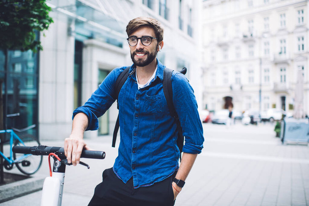 Happy young male with beard and eyeglasses in denim shirt and backpack standing with scooter on street during daylight with hand in pocket while listening to music through wireless earphones and looking away - Photo, image